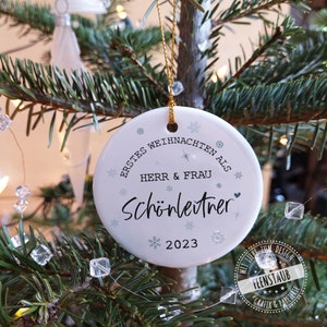 May include: White ceramic Christmas ornament with the text "Erstes Weihnachten als Herr & Frau Sch&ouml;nleitner 2023" and snowflake designs.