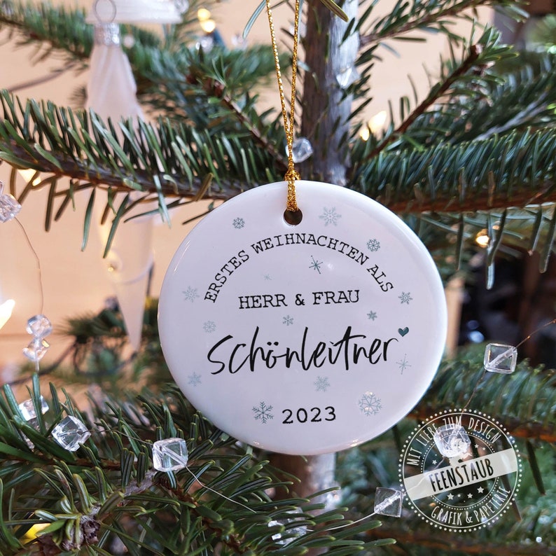 May include: White ceramic Christmas ornament with the text "Erstes Weihnachten als Herr & Frau Sch&ouml;nleitner 2023" and snowflake designs.