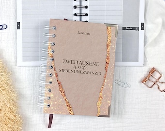 May include: A spiral-bound notebook with a light brown cover featuring the name Leonie and the text ZWEITAUSEND und SIEBENUNDZWANZIG. The cover has decorative copper-colored foil accents along the edges and metallic corner protectors.