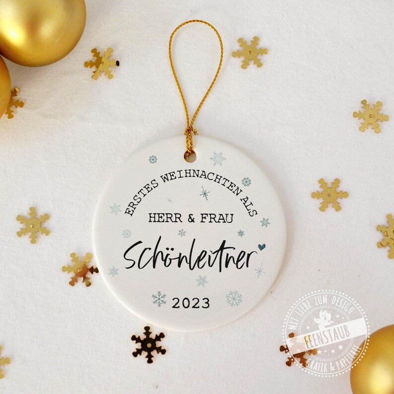 May include: White round ceramic ornament with the text "ERSTES WEIHNACHTEN ALS HERR & FRAU Sch&ouml;nleitner 2023" in black lettering with blue snowflakes and a small heart.