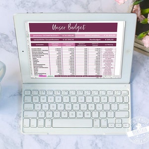 Può includere: Lo schermo di un laptop mostra un foglio di calcolo intitolato "Unser Budget" con uno schema di colori rosa e bianco. Il foglio di calcolo elenca le categorie di spese per il matrimonio, tra cui la location, il catering e i fiori, con colonne per i costi previsti, i costi effettivi e i pagamenti effettuati. Il foglio di calcolo mostra un budget totale di 16.500,00 €, costi effettivi di 16.294,50 € e un budget residuo di 205,50 €.