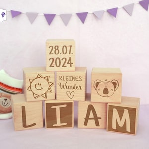 Op de afbeelding: Houten blokken die de naam "LIAM" spellen met een zon, een koala en de datum "28.07.2024" en de tekst "kleines Wunder" op andere blokken.