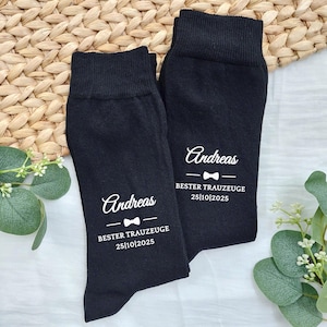 Könnte beinhalten: Zwei schwarze Socken mit weißem Text. Jede Socke hat den Namen "Andreas", eine Fliege und den Text "BESTER TRAUZEUGE 25|10|2025". Die Socken liegen auf einem gewebten Hintergrund mit Grün.