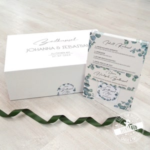 May include: A white box with the text "Zeitkapsel Johanna & Sebastian Zu Offnen Am 27-07-2023" and a sticker that says "Unsere Zeitkapsel Box".  Next to the box is a card with the text "Inhalt Kartenvel" and a list of items to include in a time capsule. The card has a eucalyptus leaf design.