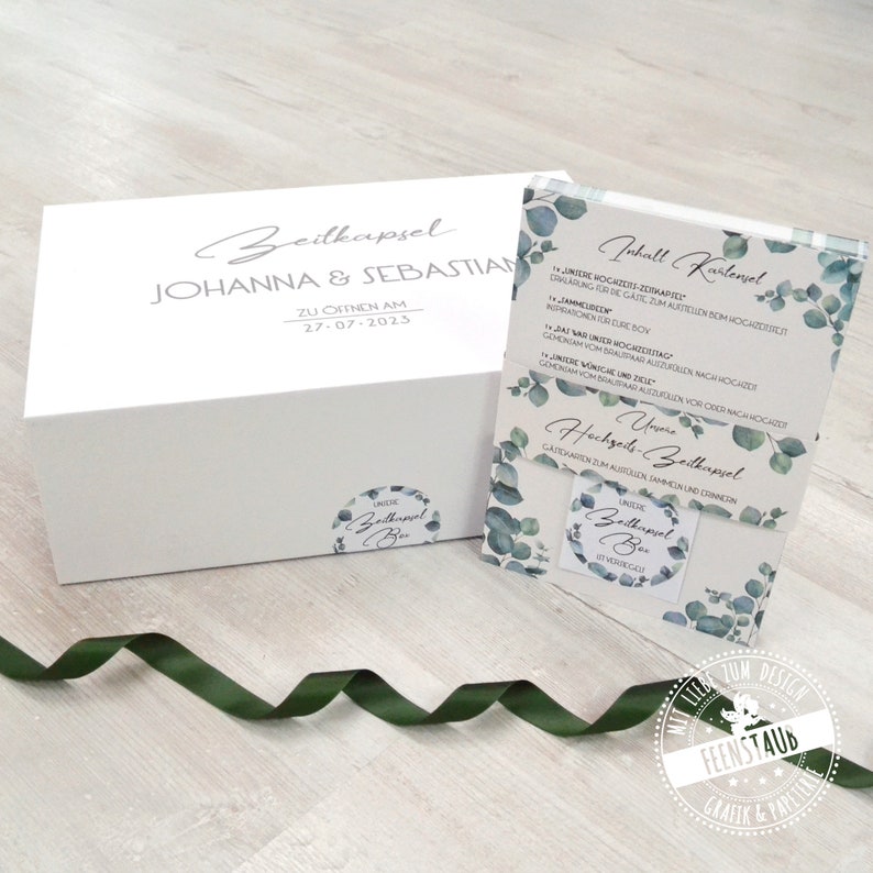 May include: A white box with the text "Zeitkapsel Johanna & Sebastian Zu Offnen Am 27-07-2023" and a sticker that says "Unsere Zeitkapsel Box".  Next to the box is a card with the text "Inhalt Kartenvel" and a list of items to include in a time capsule. The card has a eucalyptus leaf design.