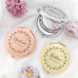 Op de afbeelding: Drie gepersonaliseerde compacte spiegels in roségoud, zilver en goud. Elke spiegel heeft een bloemenkransontwerp met een naam en de tekst "Beste Trauzeugin" in zwart schrift. De spiegels zijn open en onthullen een reflecterend oppervlak.