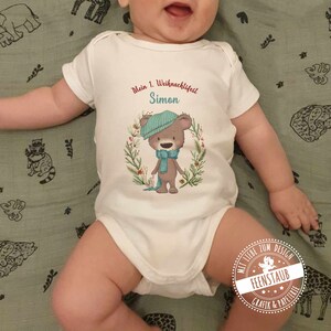 Può includere: Un body bianco per neonato con un orso marrone che indossa un cappello e una sciarpa blu. Il body ha il testo "Mein 1. Weihnachtsfest Simon" stampato su di esso.