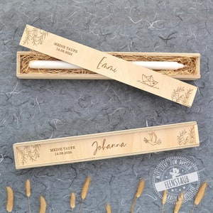 Op de afbeelding: Twee houten dozen met gegraveerde bloemmotieven en namen. De bovenste doos bevat een witte kaars en de tekst "MEINE TAUFE 14.08.2024 Emmi". De onderste doos heeft de tekst "MEINE TAUFE 14.08.2024 Johanna". Een klein logo met de tekst "MIT LIEBE ZUM DESIGN FEENSTAUB GRAFIK & LAPETERIL" is zichtbaar in de rechterbenedenhoek.
