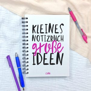 Könnte beinhalten: Ein weißes Notizbuch mit Spiralbindung und dem schwarzen sowie pinken Text KLEINES NOTIZBUCH große IDEEN und dem Namen LIANA am unteren Rand. Das Notizbuch wird von drei Stiften in Pink, Blau und Lila begleitet, die um das Objekt angeordnet sind.