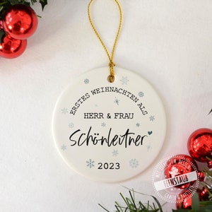 May include: White ceramic Christmas ornament with the text "ERSTES WEIHNACHTEN ALS HERR & FRAU Sch&ouml;nleitner 2023" in black and blue lettering. The ornament is decorated with snowflakes and a small heart.