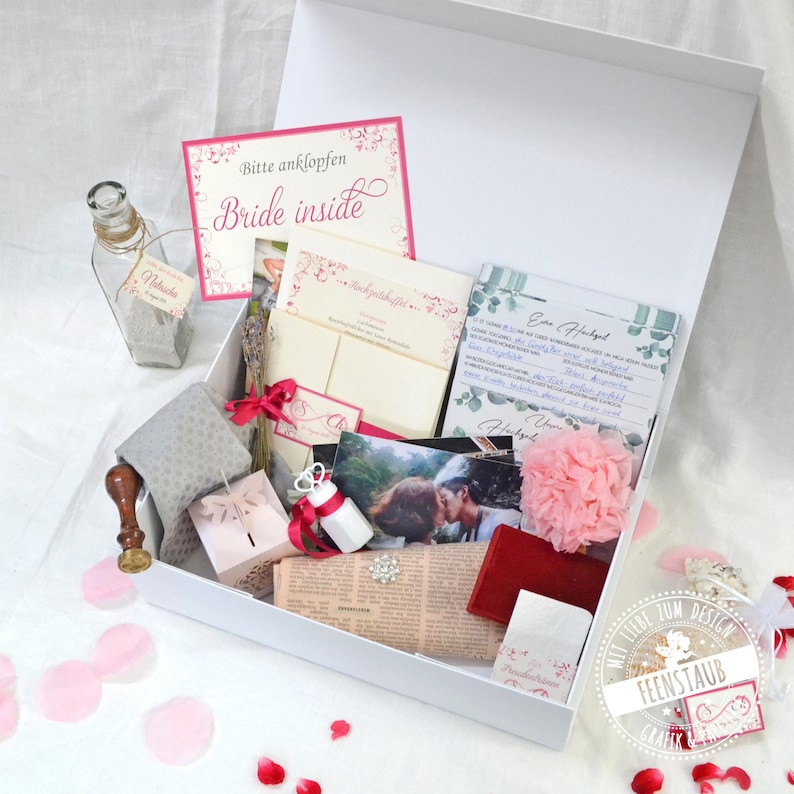 May include: A white box filled with wedding-themed items, including a bottle of sand, a small box with a butterfly design, a pink flower, a card that says "Bride Inside", and a card with the text "Ein Hochzeitsgeschenk".