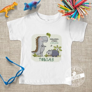 Könnte beinhalten: Weißes T-Shirt mit einem grünen Dinosaurier-Motiv und dem Text "Dinosaur-starkes Schulkind" und "Tobias".