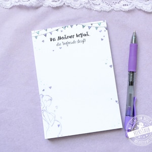 Può includere: Un blocco note bianco con un design a bandierine blu e verde e il testo "Das Abenteuer beginnt. die Vorfreude steigt!" Una donna incinta è illustrata sul blocco note. Il blocco note è su una superficie viola.