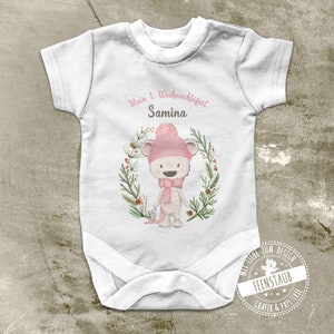 Può includere: Body bianco per neonato con un orso rosa e bianco che indossa un cappello rosa e una sciarpa. Il body ha la scritta "Mein 1. Weihnachtsfest Samina" stampata.