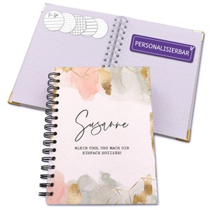 Könnte beinhalten: Ein rosa-graues Spiralnotizbuch mit goldenen Akzenten und einem Aquarelldesign. Der Einband trägt den Text "Susanne" und "BLEIB COOL UND MACH DIR EINFACH NOTIZEN!" Das Notizbuch ist mit einem violetten Aufkleber personalisiert, auf dem "PERSONALISIERBAR" steht.