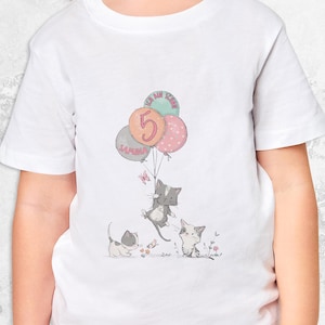Può includere: Maglietta bianca con un design di palloncini rosa, verde e arancio. I palloncini hanno il numero "5" e il nome "Samira" scritto su di essi. Due gatti dei cartoni animati stanno tenendo i palloncini. La maglietta è un ottimo regalo per una bambina di 5 anni.