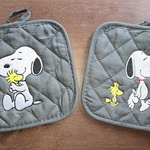 Puede incluir: Dos agarraderas grises acolchadas con diseños bordados de Snoopy y Woodstock. Una muestra a Snoopy abrazando a Woodstock, la otra a Snoopy caminando con Woodstock. Cada una tiene un lazo para colgar.