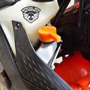 Könnte beinhalten: Nahaufnahme eines schwarzen Motorrads mit einem weißen Kunststofftank. Der Tank hat einen schwarz-weißen Aufkleber mit der Aufschrift "Special Ops Motorsports" und einem Totenkopf-Logo. Der Tank hat einen orangenen Deckel mit einem Hebel.
