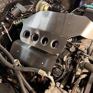 May include: Close-up of a car engine with a silver metal cover over the intake manifold. The engine is black and has several hoses and wires attached to it.