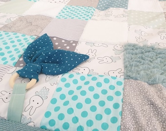 May include: A baby play mat with a variety of textures and patterns. The mat features a blue and white polka dot fabric, a gray and white striped fabric, a white fabric with bunny silhouettes, and a light blue fuzzy fabric. A blue and white polka dot fabric bow with a wooden ring is on the mat.
