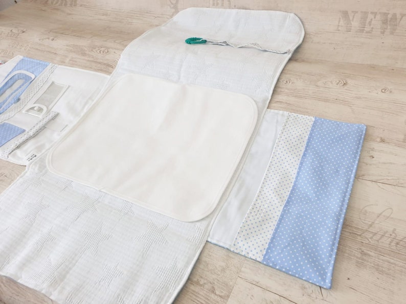 May include: A white changing mat with a blue and white patterned flap. The mat is folded in half and has a white changing pad inside. The mat is designed for changing babies and has a pocket for storing wipes or other baby essentials.