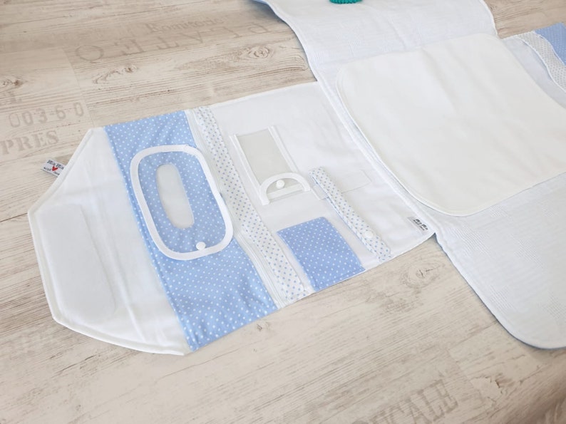 May include: A white and light blue baby changing mat with multiple pockets and compartments. The mat is folded open to reveal a clear plastic pocket, a blue polka dot pocket, and a white pocket with a white zipper. The mat is made of a soft, quilted fabric and is perfect for keeping all of your baby's essentials organized.
