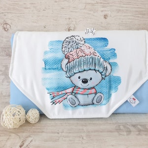 May include: A white baby burp cloth with a blue watercolor background and a cartoon bear wearing a blue and pink hat and a pink and white scarf. The burp cloth is folded in half and has a blue backing.