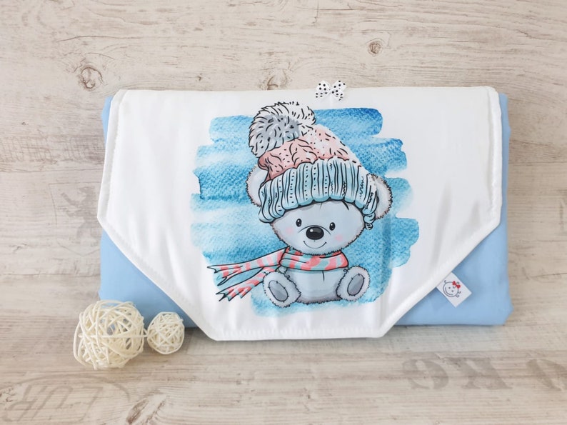 May include: A white baby burp cloth with a blue watercolor background and a cartoon bear wearing a blue and pink hat and a pink and white scarf. The burp cloth is folded in half and has a blue backing.