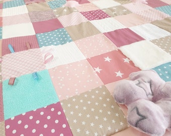 May include: A multi-colored baby play mat with various textures and patterns. The mat features pink, white, brown, and blue fabrics with different designs, including stars, polka dots, and geometric shapes. A plush purple toy is on the mat.