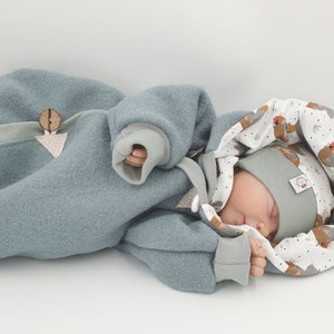 Peut inclure: Un bébé portant une combinaison à capuche bleu clair avec un détail étoile et bouton en bois. La capuche est doublée d'un tissu blanc à motif d'oiseaux brun et blanc. Le bébé porte un bonnet bleu clair assorti.