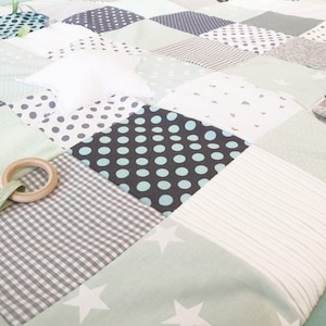 May include: A baby play mat with a patchwork design featuring various textures and patterns. The mat includes a wooden ring, a white star, and a green ribbon.