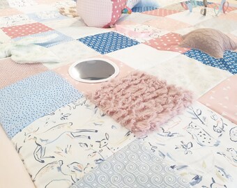 May include: A multi-colored patchwork play mat with various textures, including a mirror, fur, and a knitted pillow. The mat is made of different fabrics in shades of pink, blue, and white.