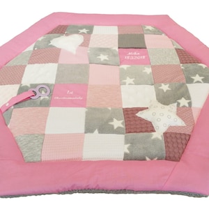 Puede incluir: Alfombra de juego de patchwork rosa y gris con un diseño de estrellas y corazones. La alfombra tiene un borde rosa y una etiqueta blanca con el texto "1er Bebé de Navidad".