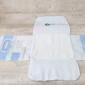 May include: A white changing pad with a blue and white patterned border. The changing pad is folded in half and has a small green clip attached to it.