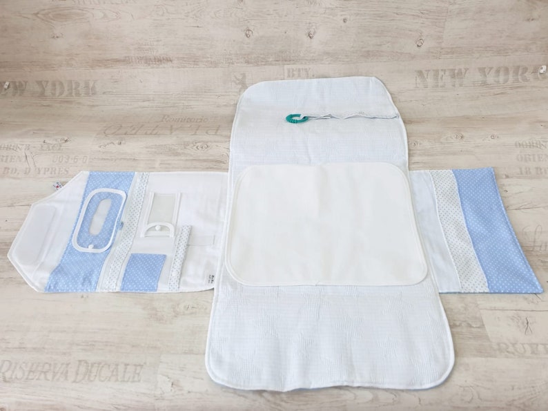 May include: A white changing pad with a blue and white patterned border. The changing pad is folded in half and has a small green clip attached to it.