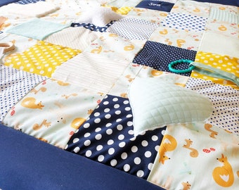 May include: A multi-colored patchwork baby play mat with a variety of textures. The mat features a blue border, yellow and white polka dot fabric, a white fabric with a striped pattern, and a white fabric with a fox print. The mat also includes a light blue pillow and a green teething ring.
