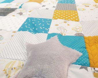 Peut inclure: Une courtepointe bébé patchwork avec une variété de tissus, dont un tissu bleu avec des triangles blancs, un tissu blanc avec des pois gris, un tissu jaune avec des pois blancs et un tissu gris avec un motif en tricot torsadé. Un oreiller en forme d'étoile gris se trouve au premier plan.