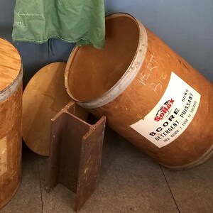 Pode incluir: Um armário de madeira com prateleiras, um avental verde pendurado em um gancho, um martelo e dois barris de madeira. Um barril está etiquetado "Score Detergente Puissant 20 kg" e o outro está etiquetado "Vert".