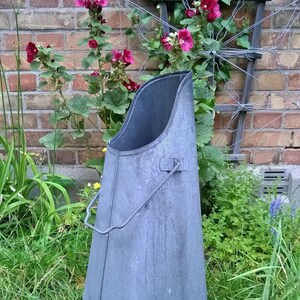Peut inclure: Seau à charbon en métal gris, avec une poignée, posé dans l'herbe. Le seau a une large ouverture et une forme conique. En arrière-plan, un mur de briques et des plantes à fleurs. Idéal pour le jardin.