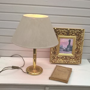 Könnte beinhalten: Ein weißer Schrank mit Glastüren und einer weißen Oberseite. Eine Lampe mit einem beigefarbenen Lampenschirm und einem goldenen Fuß steht auf der Oberseite des Schranks. Ein kleines gerahmtes Bild mit einem goldenen Rahmen steht auf der Oberseite des Schranks. Ein Buch liegt auf der Oberseite des Schranks.