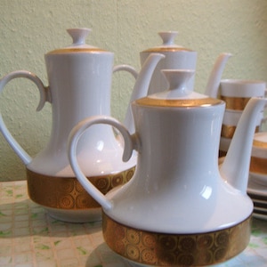 Może przedstawiać: Zestaw białych porcelanowych czajników i filiżanek z pozłacanymi zdobieniami i misternymi wzorami. Czajniki mają klasyczny design z zaokrąglonym korpusem i dzióbkiem. Filiżanki są małe i delikatne, z uchwytem po boku.