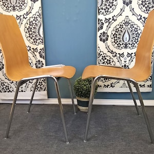 May include: Two wooden chairs with chrome legs and a simple design. The chairs are in front of a wall with black and white patterned wallpaper.