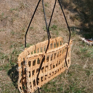 Könnte beinhalten: Ein geflochtener Weidenkorb mit einem schwarzen Metallrahmen. Der Korb ist rechteckig und hat einen Griff oben. Der Korb steht auf einem Bett aus trockenem Gras.