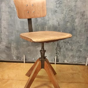 Peut inclure: Chaise en bois vintage avec assise et dossier brun clair. Le dossier est fixé à un support métallique avec quatre rivets visibles. La chaise a une base tripode en bois et un mécanisme réglable en métal.