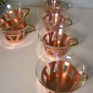 May include: Set of six clear glass cups with copper colored bands and gold handles. The cups have a geometric pattern in the copper bands.