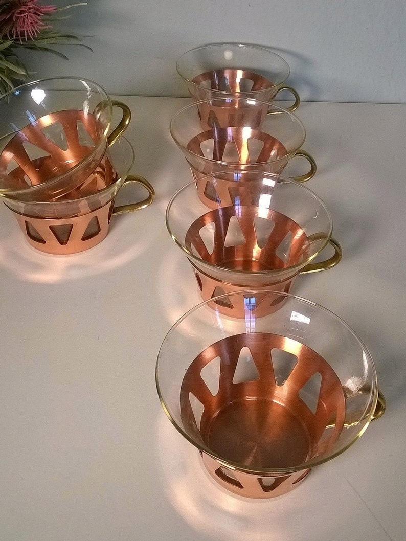 May include: Set of six clear glass cups with copper colored bands and gold handles. The cups have a geometric pattern in the copper bands.