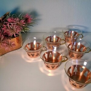 May include: Set of six clear glass teacups with copper colored metal bases. The cups have a geometric design and gold colored handles.