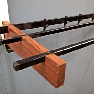 May include: A wall-mounted coat rack with a wooden base and black metal bars. The rack features a rectangular wooden beam with a natural wood grain, intersected by horizontal black metal bars with hooks for hanging items. The design is minimalist and functional.