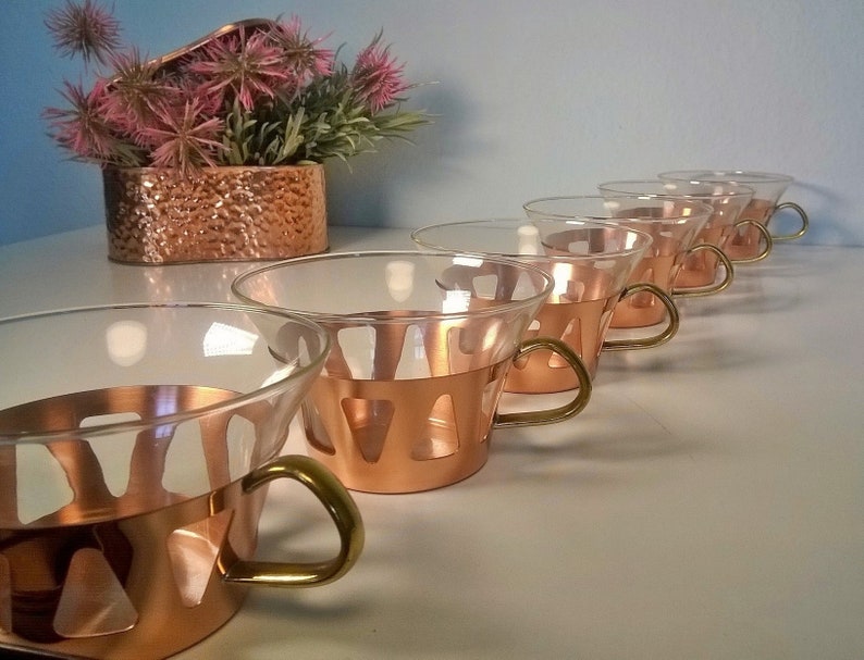 May include: Set of six copper and glass cups with brass handles. The cups have a unique design with cutouts in the copper band.
