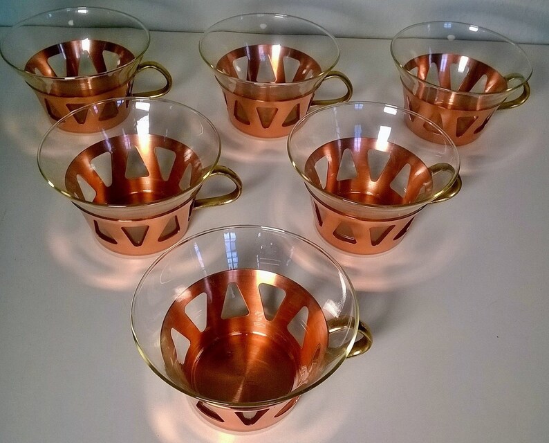 May include: Set of six copper and glass teacups with a geometric design. The cups have a clear glass bowl with a copper band around the top and a copper handle.